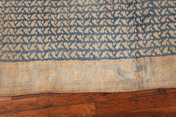 Close-up of a shaggy blue and tan geometric rug border lying on dark reddish-brown wood planks.