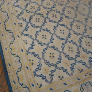 Beige high-pile rug with blue geometric lattice and yellow motifs on wood plank flooring.