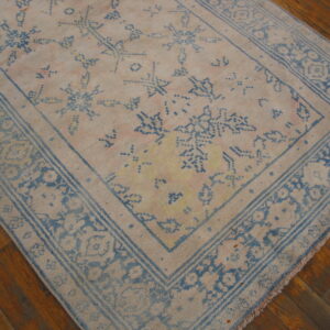 Distressed pale beige rug with blue abstract motifs and floral borders rests on rich brown wood flooring.