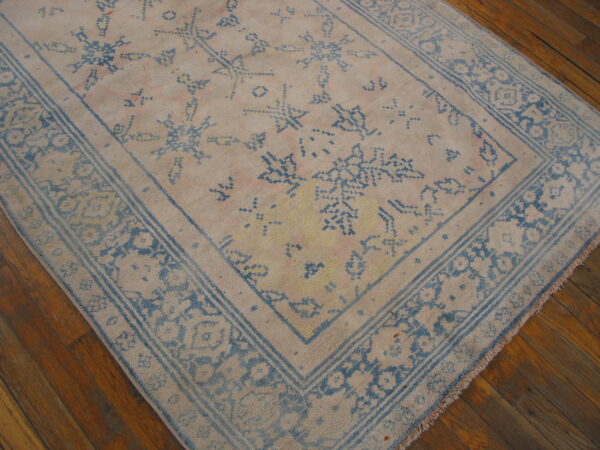 Distressed pale beige rug with blue abstract motifs and floral borders rests on rich brown wood flooring.
