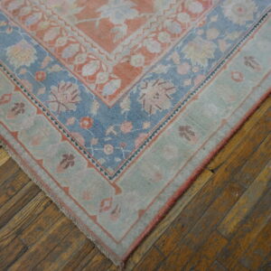 Low-pile rug corner with peach, pale blue, and sage floral patterns lying on worn hardwood planks.