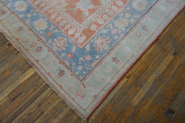 Low-pile rug corner with peach, pale blue, and sage floral patterns lying on worn hardwood planks.