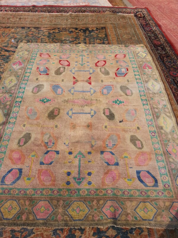 Faded pink and tan rug featuring brightly colored geometric and abstract motifs layered on other rugs.