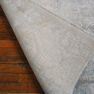 A light blue rug corner is lifted to reveal its woven backing resting on reddish-brown floors.