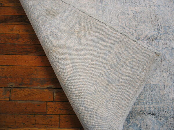 A light blue rug corner is lifted to reveal its woven backing resting on reddish-brown floors.