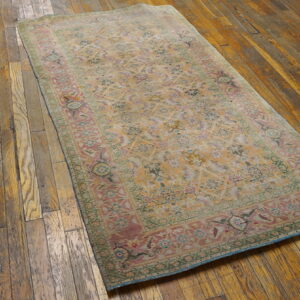 Faded traditional rug in gold, pink, and green lies diagonally on a distressed dark wood floor.