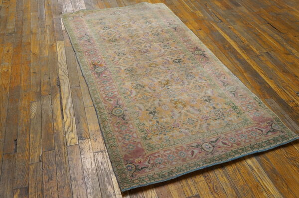 Faded traditional rug in gold, pink, and green lies diagonally on a distressed dark wood floor.