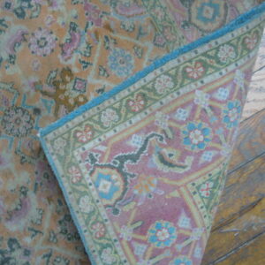 A low-pile orange and pink floral rug corner is lifted, showing the woven pink, green, and blue backing on wood floors.
