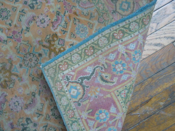 A low-pile orange and pink floral rug corner is lifted, showing the woven pink, green, and blue backing on wood floors.