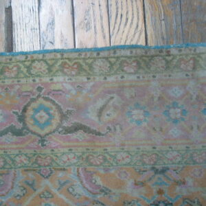 Muted floral patterned rug in pink and green with a blue edge against rustic hardwood flooring.