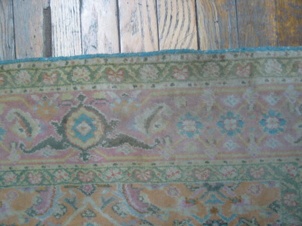 Muted floral patterned rug in pink and green with a blue edge against rustic hardwood flooring.