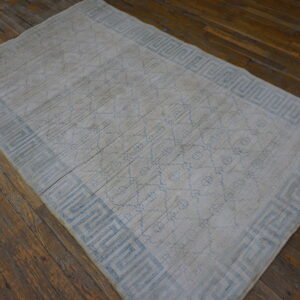 Low-pile white rug with a light blue meander border pattern on distressed dark hardwood flooring.