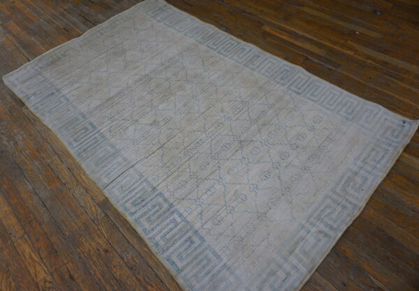 Low-pile white rug with a light blue meander border pattern on distressed dark hardwood flooring.