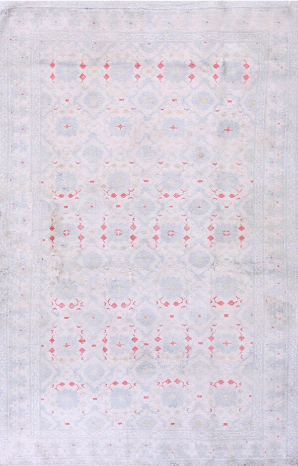 Low pile rug in faded pale blue, pink, and white geometric patterns on distressed wood flooring.