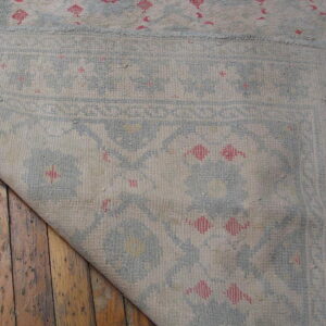 Light blue and beige rug with pink details folded back over distressed wood plank flooring.