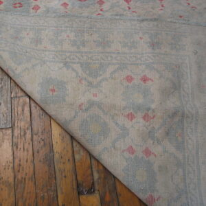 Light beige rug with blue and pink motifs folded back, revealing distressed wood floorboards.