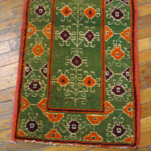 High-pile green rug featuring bold orange and purple geometric designs rests on distressed hardwood flooring.