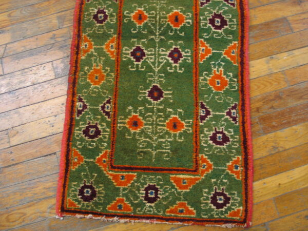 High-pile green rug featuring bold orange and purple geometric designs rests on distressed hardwood flooring.