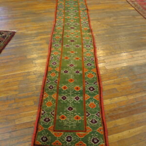 Green runner with orange and purple geometric patterns and red edging on worn wooden plank flooring.