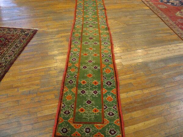 Green runner with orange and purple geometric patterns and red edging on worn wooden plank flooring.