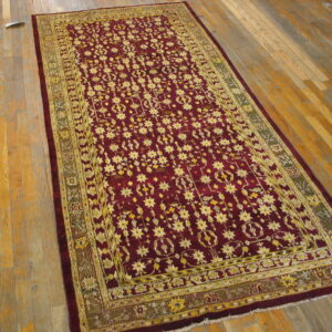 Burgundy rug with a yellow repeating floral pattern and olive green border on worn wood planks.