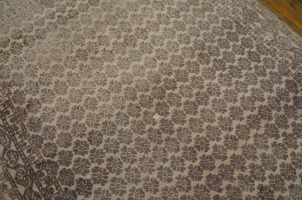 Low-pile taupe rug with repeating dark floral motifs, a patterned border, resting on wood flooring.