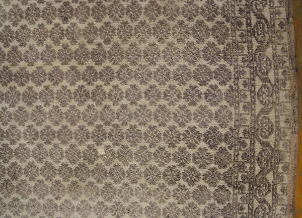 Detail shot of a beige rug patterned with repeating dark taupe floral clusters and a textured border.