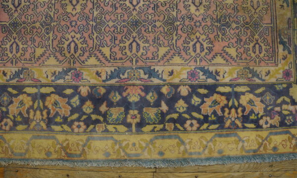 Detail of a traditional patterned rug featuring rose, indigo, and gold borders, resting on wood floorboards.