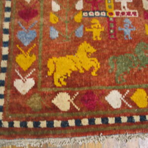 Rust-red rug with multicolored folk art motifs, including hearts and horses, showing a blue edge and fringe on wood.