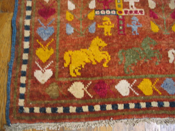 Rust-red rug with multicolored folk art motifs, including hearts and horses, showing a blue edge and fringe on wood.