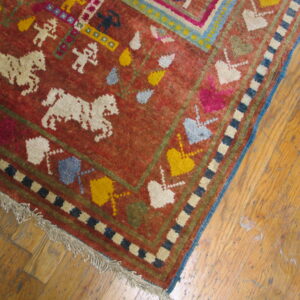 Low-pile red rug with colorful figures, horses, and geometric borders rests on worn wood flooring.