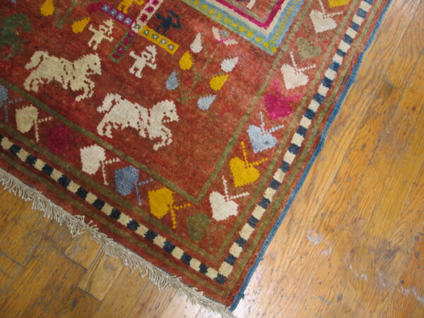 Low-pile red rug with colorful figures, horses, and geometric borders rests on worn wood flooring.