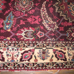 Detail of a low-pile rug with a burgundy field and cream border displaying stylized floral motifs on wood flooring.