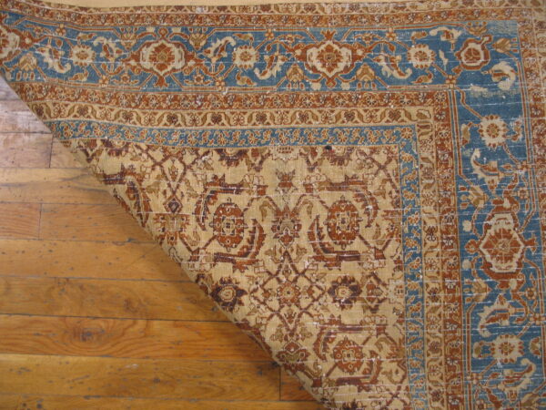 A patterned beige, rust, and blue rug is curled back over distressed wood flooring.
