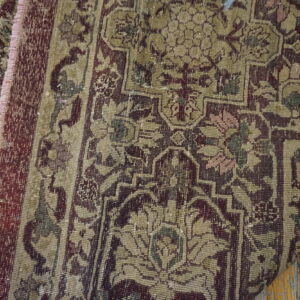 Partially folded, distressed traditional rug in muted burgundy and khaki lies on a wooden floor.