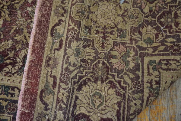 Partially folded, distressed traditional rug in muted burgundy and khaki lies on a wooden floor.