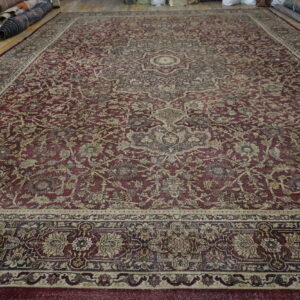 Large red and beige traditional rug displayed on wood floor with other rolled rugs nearby.