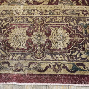 Ornate low-pile rug border in tan, deep red, and dark green, placed on light gray wood flooring.