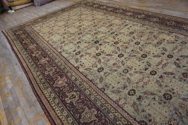 Oversized distressed rug featuring a light sage field and intricate burgundy and cream patterned border on wood floorboards.