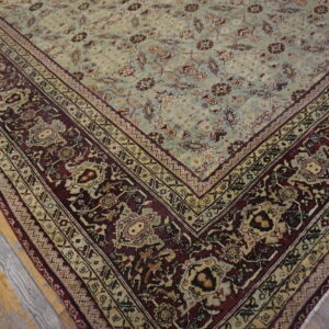 Muted green central field rug with elaborate maroon and beige borders rests on weathered hardwood flooring.