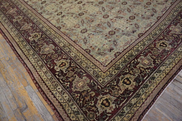 Muted green central field rug with elaborate maroon and beige borders rests on weathered hardwood flooring.
