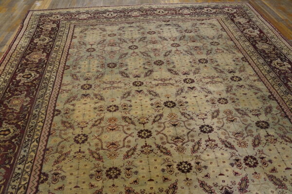 Traditional patterned rug featuring a light greenish-beige field and wide maroon borders on a wood plank floor.