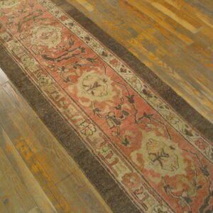 Muted coral and tan ornamental runner rug featuring a dark brown border on distressed wood plank flooring.