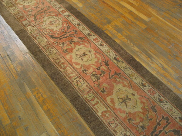 Muted coral and tan ornamental runner rug featuring a dark brown border on distressed wood plank flooring.