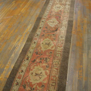 A coral and tan patterned runner rug with a wide dark border lays diagonally on worn wood planks.