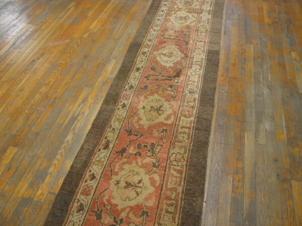 A coral and tan patterned runner rug with a wide dark border lays diagonally on worn wood planks.