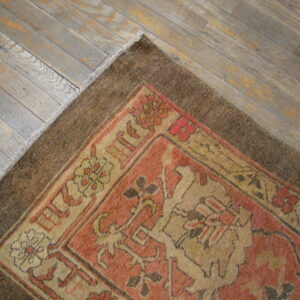 Muted terracotta and tan patterned rug with a brown border rests on gray-toned wood flooring.