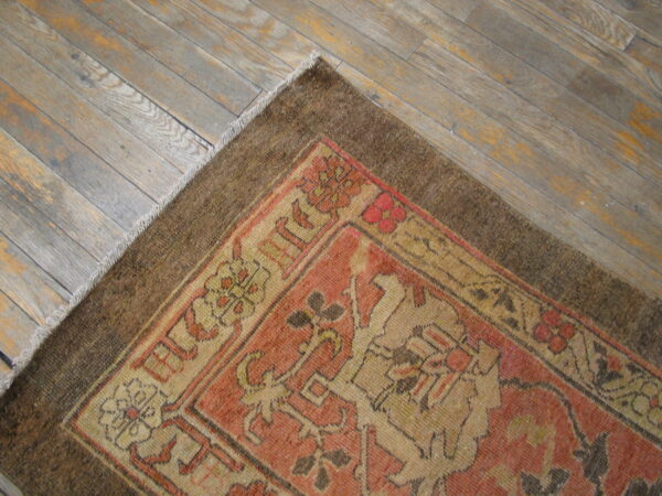 Muted terracotta and tan patterned rug with a brown border rests on gray-toned wood flooring.