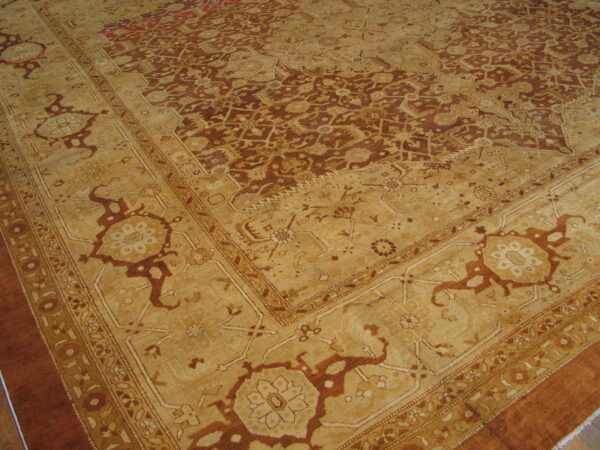 Angled view of a low-pile traditional rug with dense red-brown and tan geometric patterns and a detailed border.