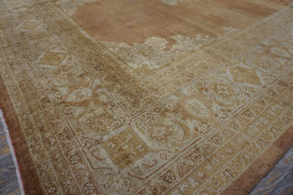 Ornate beige and brown traditional border detail of a low pile rug lying on gray wood flooring.
